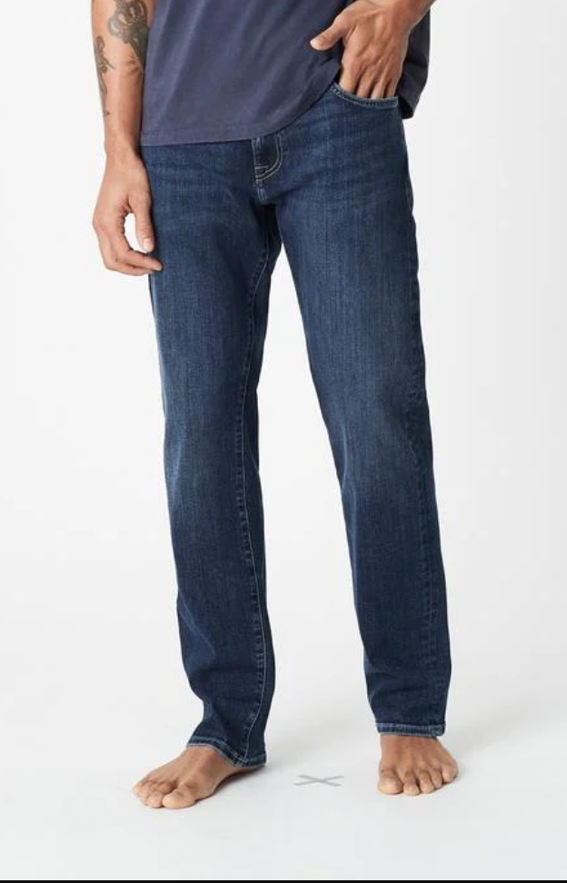 Barefoot, a person in a dark blue t-shirt showcases Mavi Zach Straight Jean in indigo blue, with relaxed fit jeans. One hand rests in the pocket, hinting at tattoos on the arm. The organic cotton fabric offers comfort against the plain white background.
