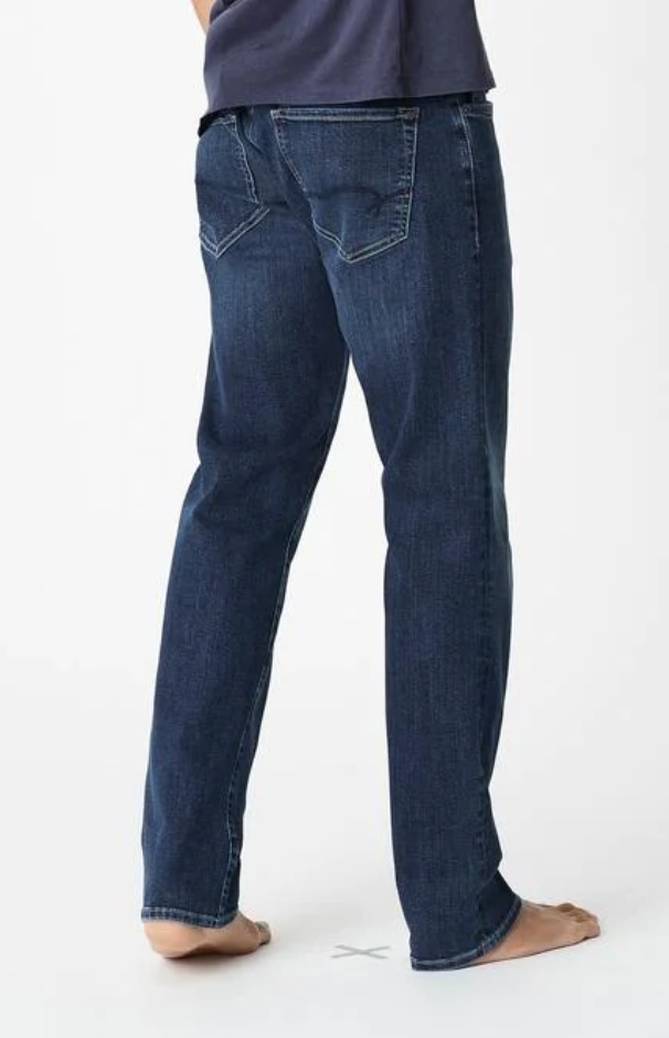 A person stands on a white background, in Mavi Zach Straight Jeans and a dark shirt. The loose-fitting, slightly faded jeans are made of organic cotton with distinct stitching on the back pockets. They are barefoot, adding to the jeans' effortless charm.