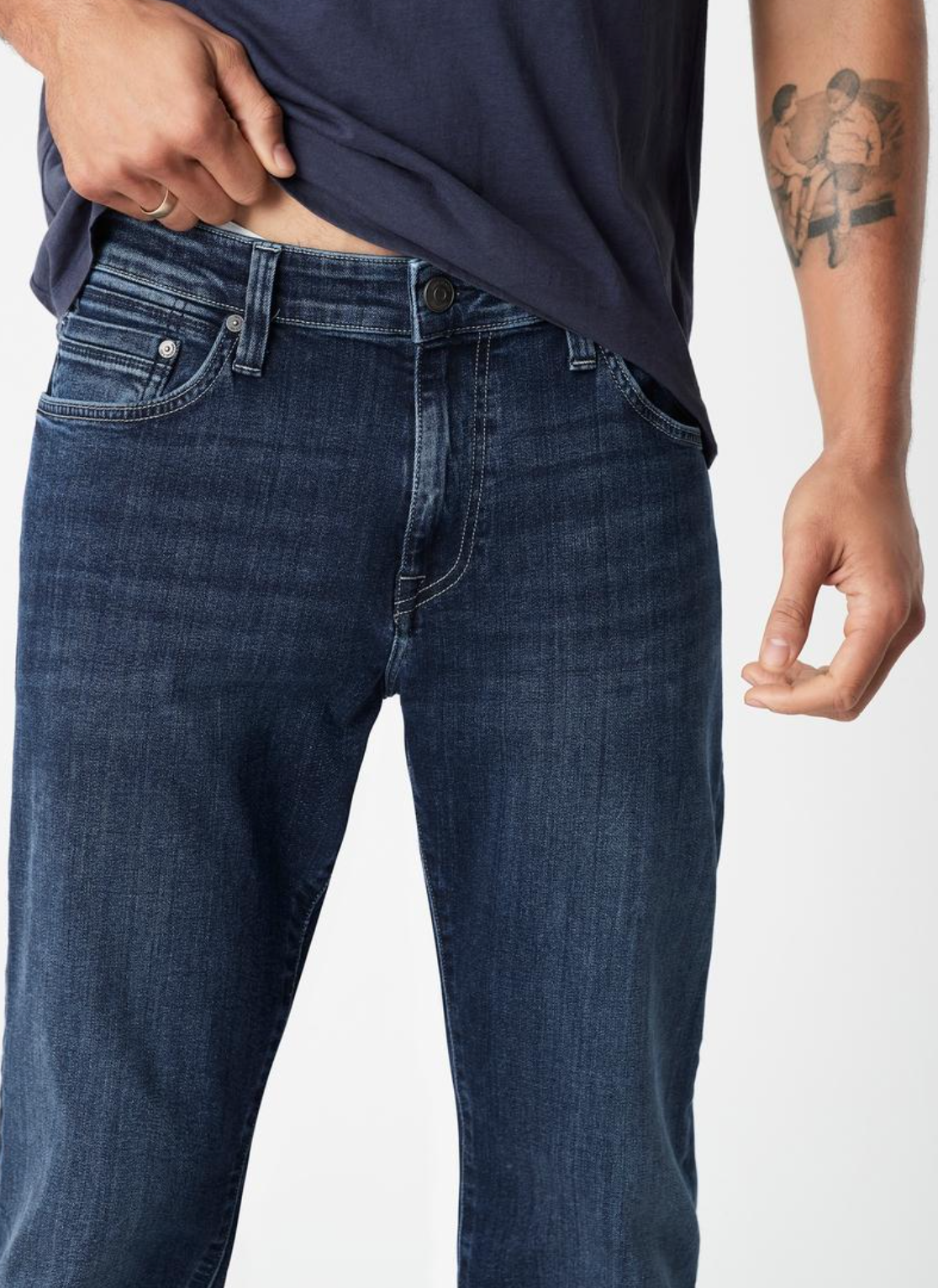Someone is wearing a navy blue shirt and Mavi's Zach Straight Jean in dark blue. They're adjusting their shirt near the waistband with their right hand, partially revealing a tattoo of two figures on their left arm against a plain white background.
