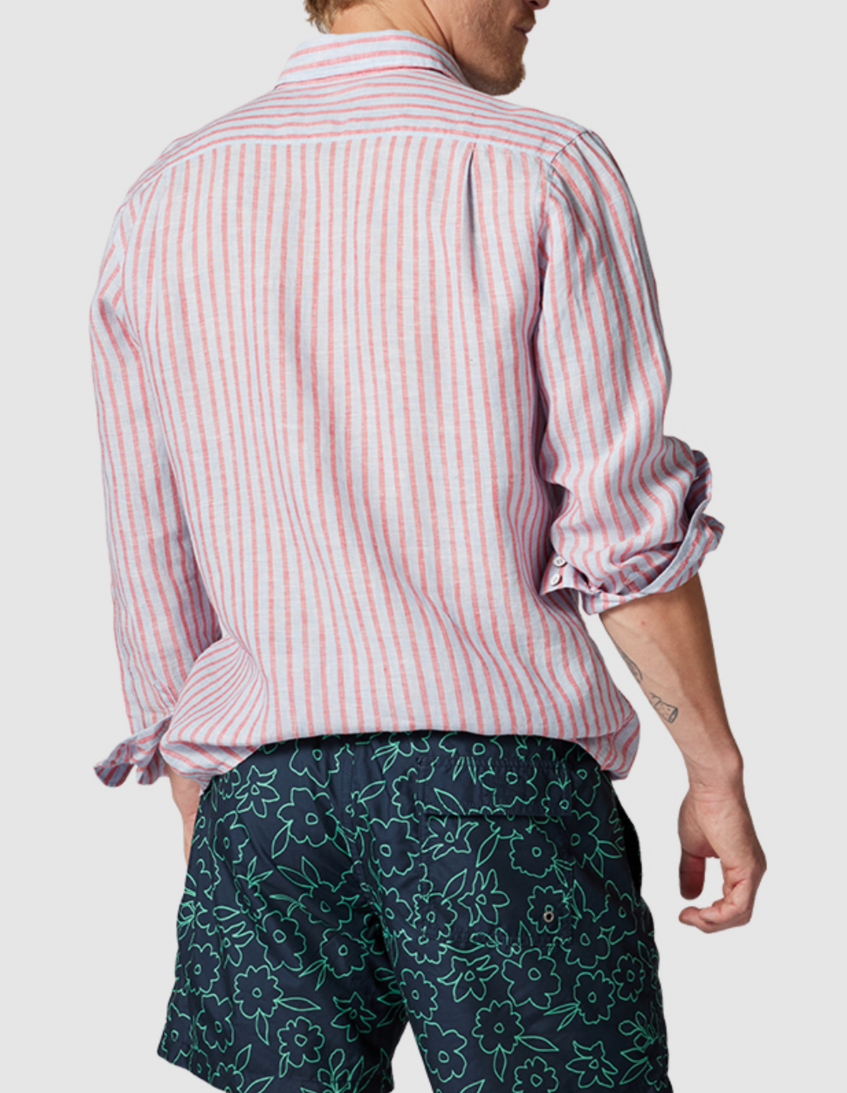 With their back to the camera, they wear a 100% linen pink and white striped Rodd & Gunn Mclean Park Oxford Sports Fit Shirt with rolled-up sleeves, paired effortlessly with navy shorts featuring a green floral pattern. The backdrop is a simple light gray.