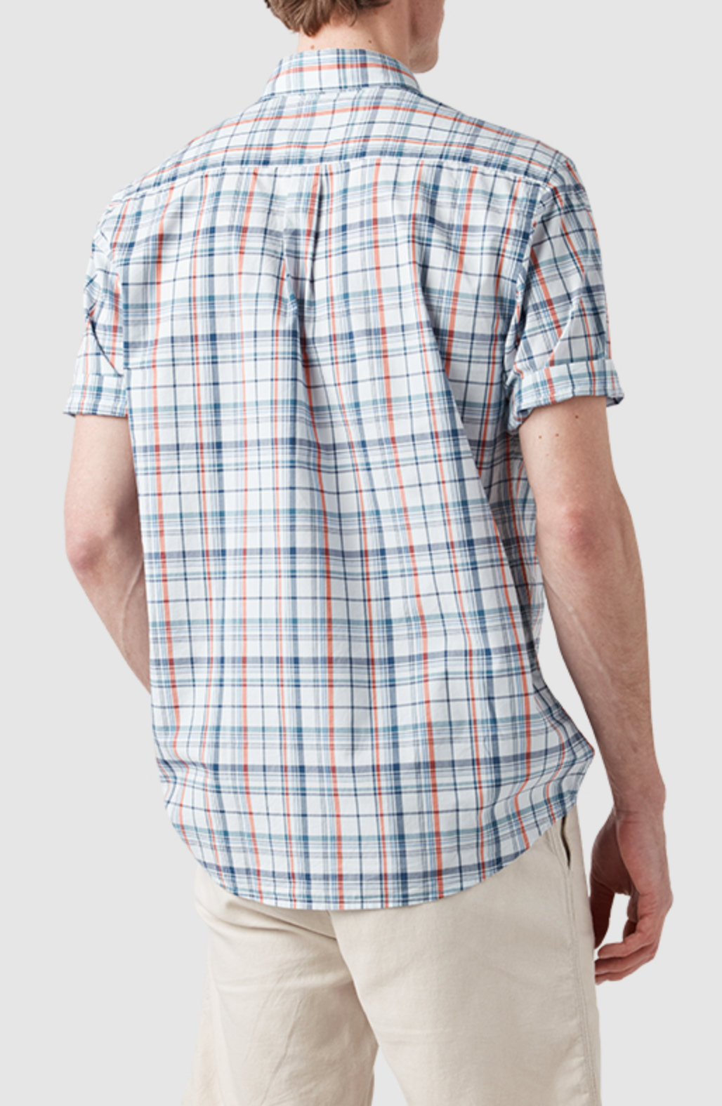 A person wearing a short-sleeved, original fit Rodd & Gunn Rodeo Drive Shirt in white and blue plaid with red accents, paired with beige pants, faces away from the camera against a gray background. The shirt is 100% cotton.
