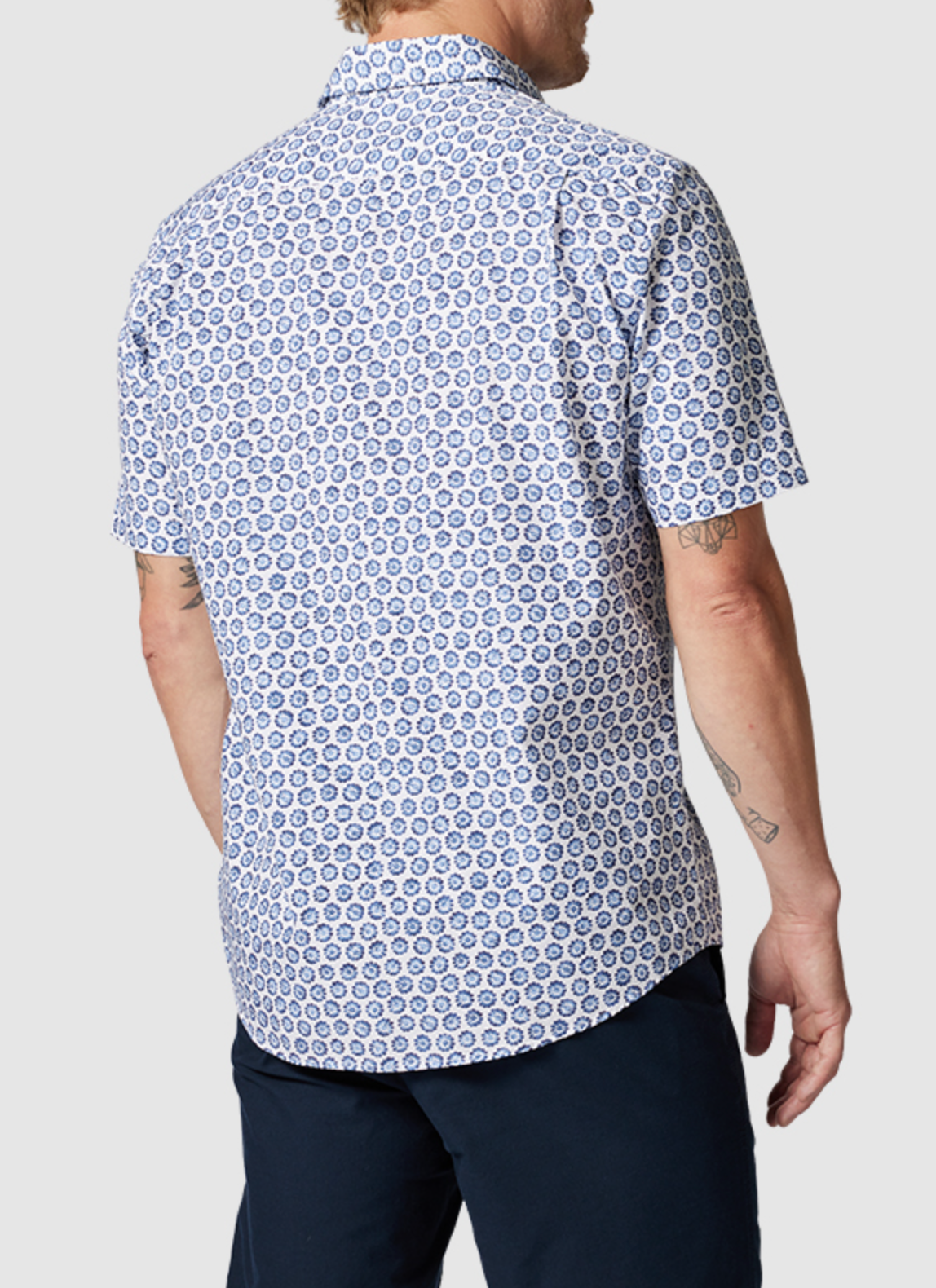 The individual sports a short-sleeved, button-up Rodd & Gunn First Avenue Shirt featuring a blue and white circular pattern and spread collar. Made of 100% cotton, it complements their dark pants. Their arm tattoos stand out against the plain gray backdrop from behind.