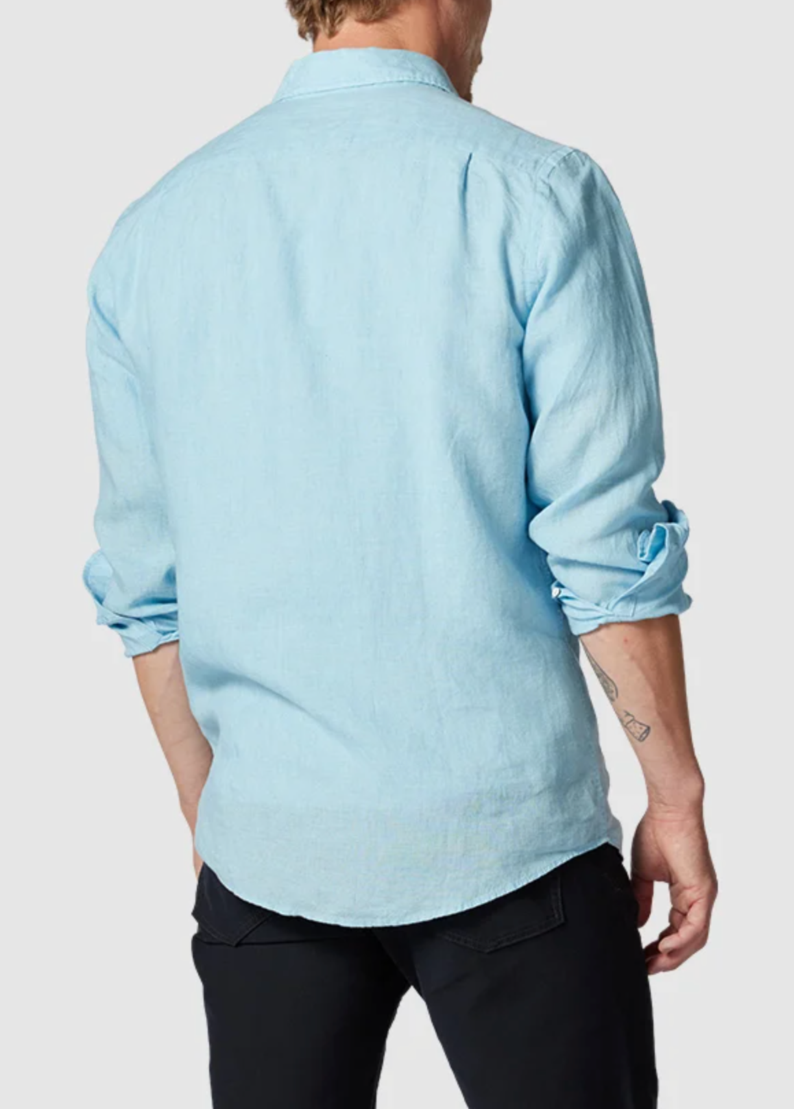 A person with sandy hair is seen from the back, wearing a light blue Rodd & Gunn Coromandel Sports Fit Shirt paired with black pants. The sleeves are rolled up to reveal a small tattoo on the right forearm, set against a plain light gray background.