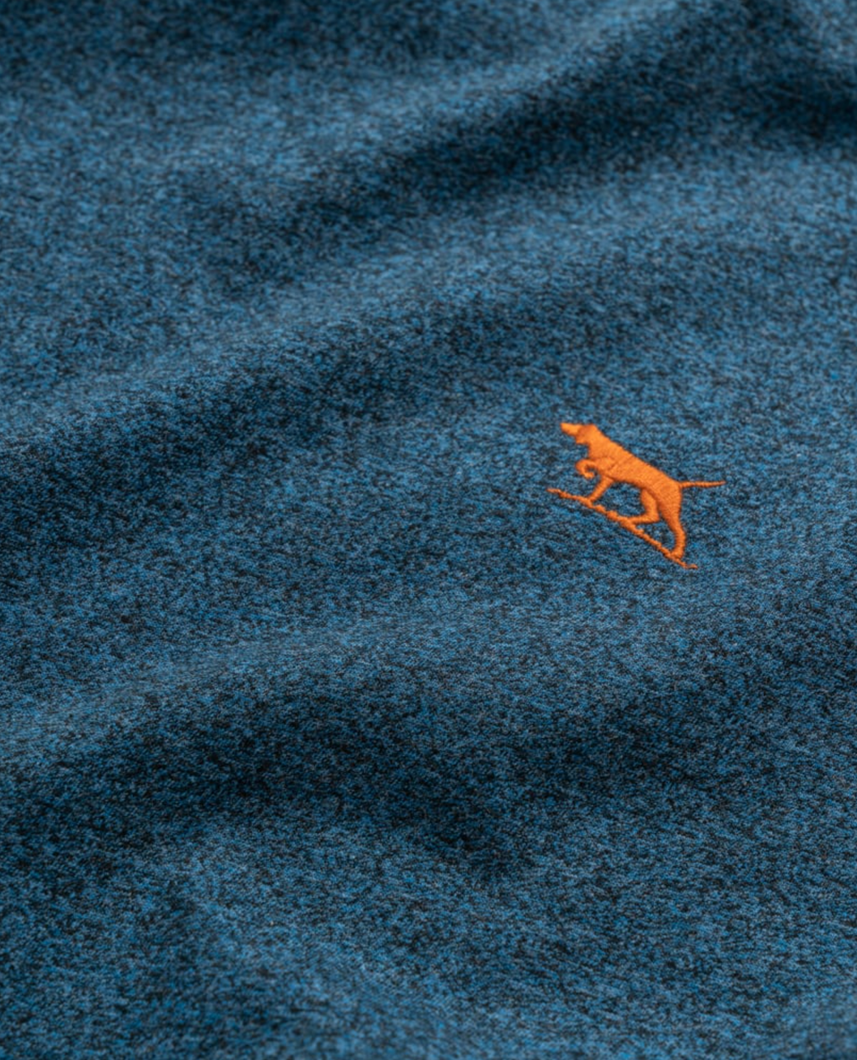 A close-up of the Rodd & Gunn The Gunn T-Shirt in blue Australian Cotton shows a textured look with an embroidered design of an orange dog holding a stick, providing a playful contrast to the fabric's hue.