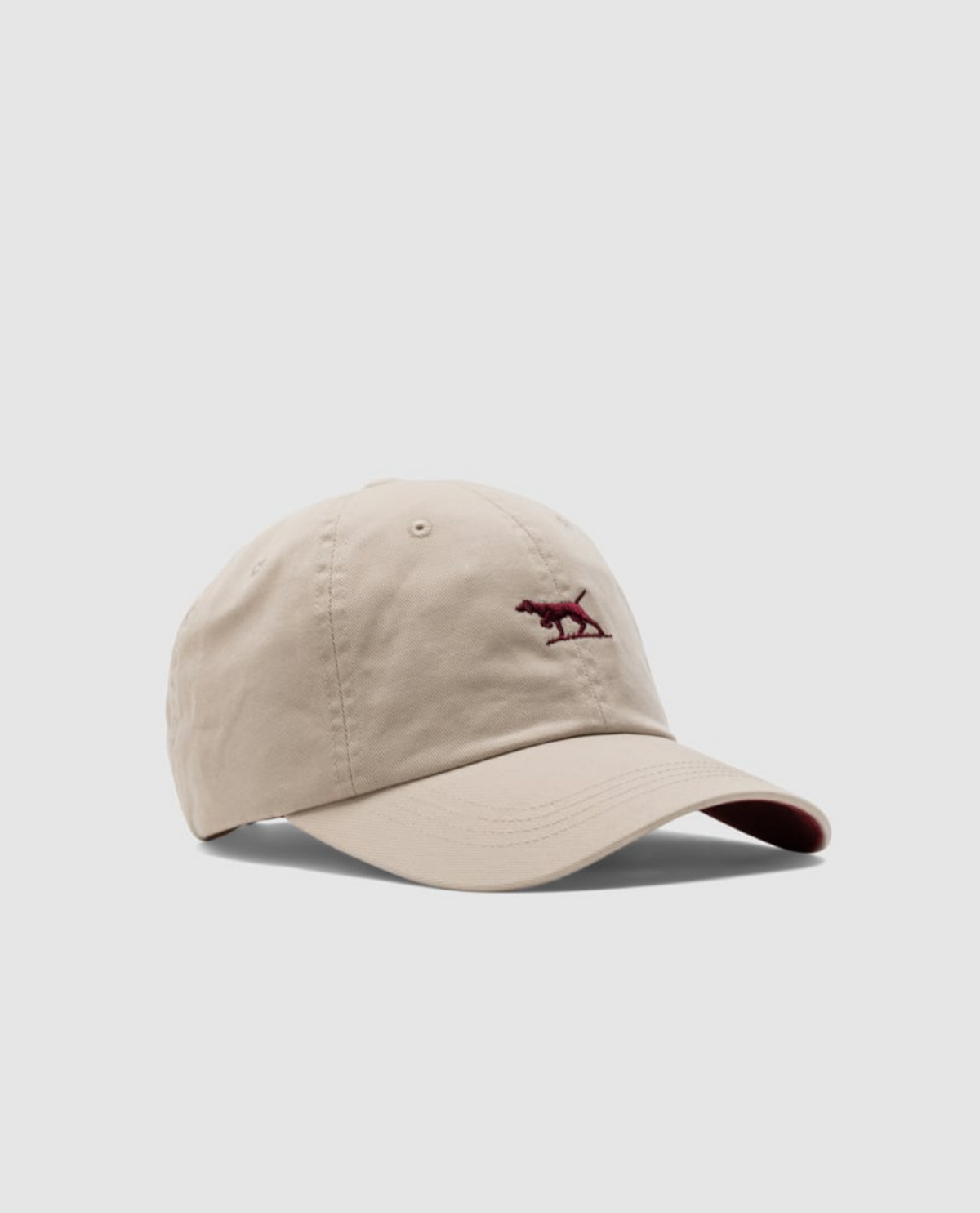 The beige Gunn Cap from Rodd & Gunn features a curved brim and durable cotton twill, with a small red embroidered animal logo on the front against a plain gray background.