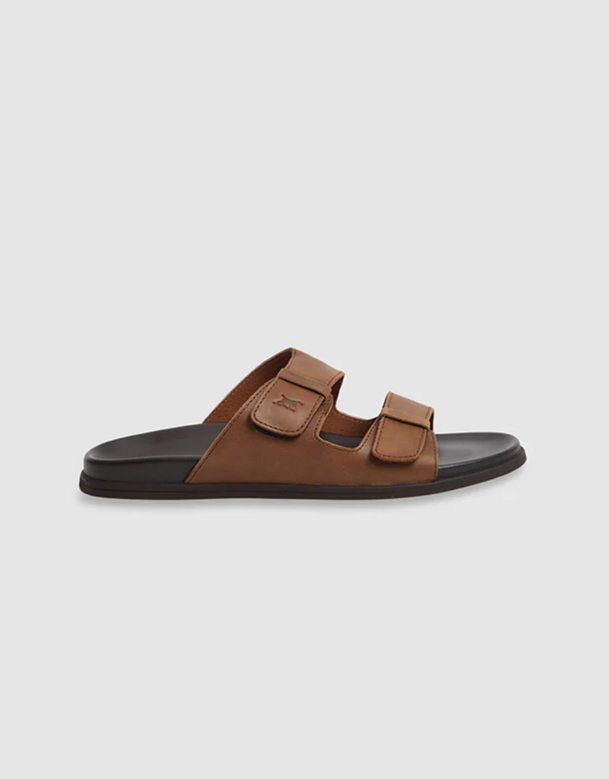 Rodd & Gunn Kendrick Place Slide sandals feature two adjustable brown leather straps, a black sole with a molded footbed for comfort, and a flexible rubber sole for easy movement. The minimalist design is showcased on a plain white background.