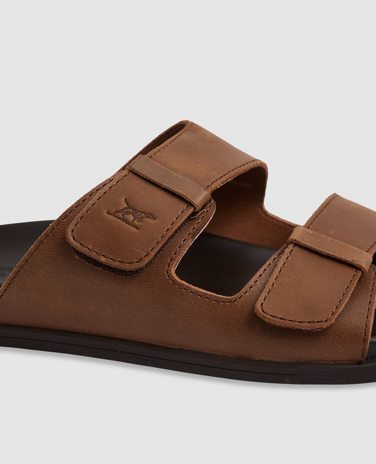 Close-up of a Rodd & Gunn Kendrick Place Slide, featuring brown leather straps with two adjustable buckles, a moulded footbed, and a flexible rubber sole. Visible stitching adorns the edges, with a small animal logo embossed on the strap against a plain white background.