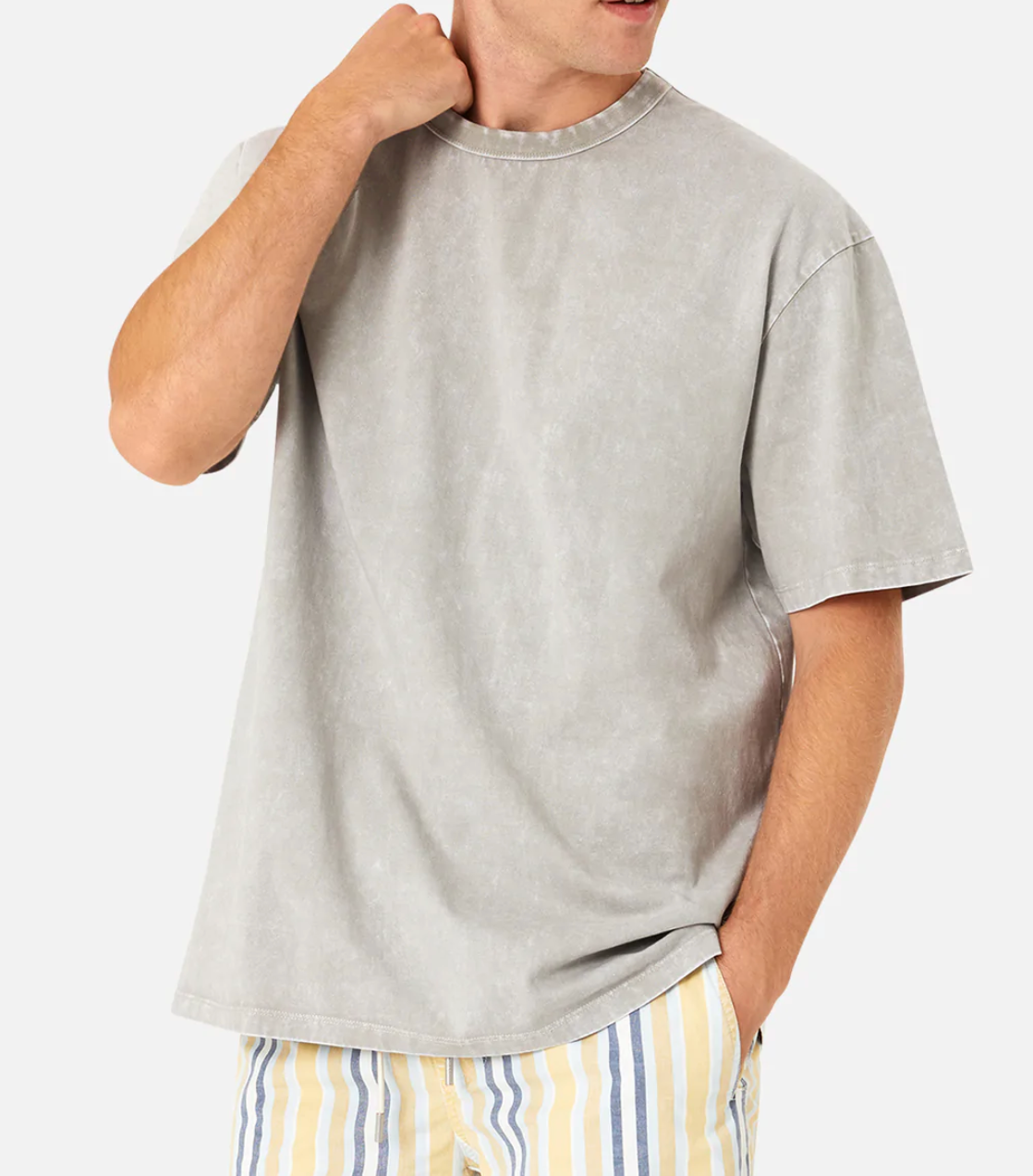 Dressed in an oversized Industrie The Del Sur Acid Wash Tee in light gray and yellow-gray striped shorts, a person stands casually against a plain white background, one hand in their pocket and the other touching their neck.