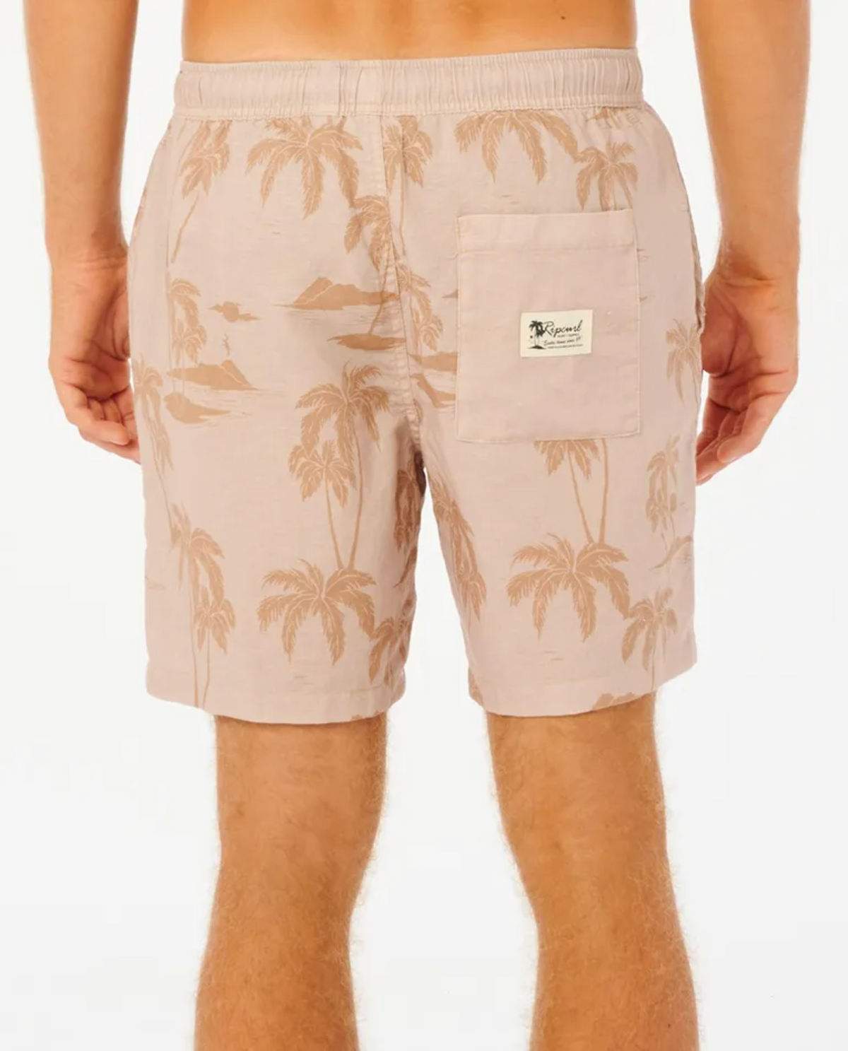 A person is seen from behind wearing Rip Curl Mod Tropics Volley shorts with a tropical palm tree design. These linen/cotton blend shorts feature an elastic waist, a back pocket with a small white label, and reveal the skin on the legs.