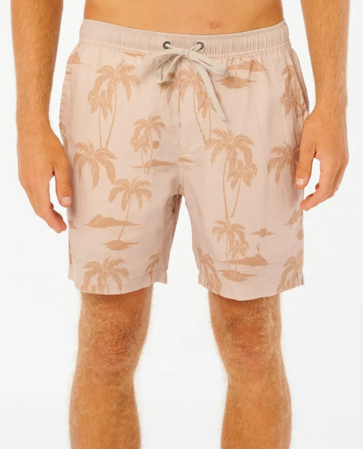 A person wears Rip Curl Mod Tropics Volley shorts in light beige with a palm tree and island pattern. Made from a premium linen/cotton blend, these fashionable shorts feature an elastic drawstring waistband. The white background highlights the person's legs and hands.