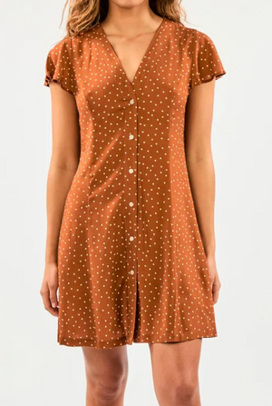 A person stands against a white background, wearing the Rip Curl Dreamer Spot Button Through Dress, a relaxed-fit dress in a brown EcoVero/Viscose blend with small white polka dots and a V-neckline.