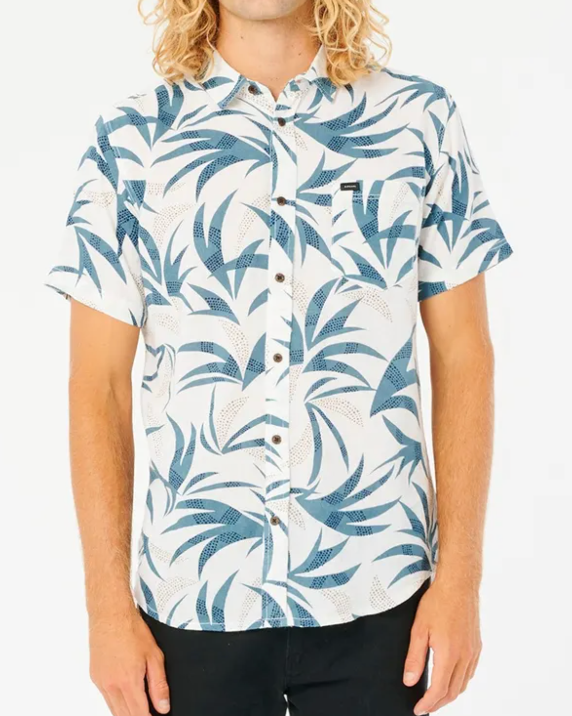 The person wears a Rip Curl Angourie Short Sleeve Shirt with a blue and white leaf pattern, paired with black pants. Their long, curly blonde hair cascades as they stand against a plain white background.