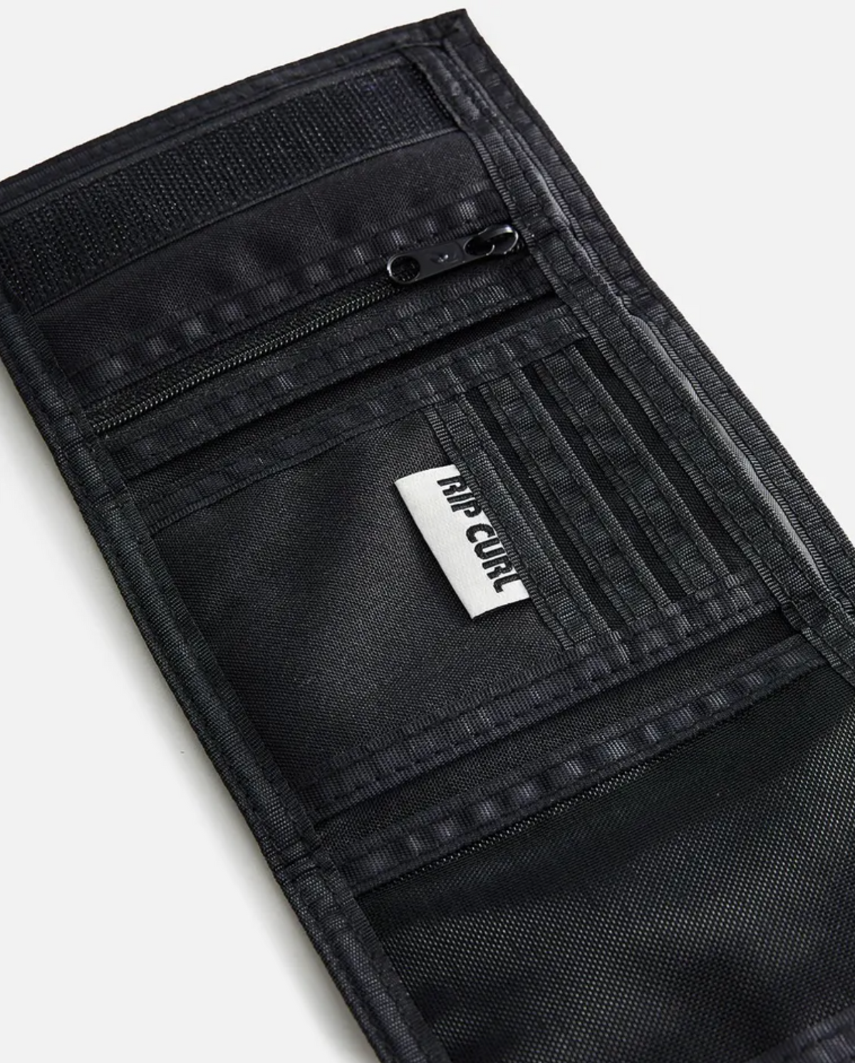 A close-up of the Rip Curl Surf Revival Surf Wallet, an open black recycled polyester wallet featuring multiple compartments, a zippered pocket, card slots, and a section with a white tag displaying black text.