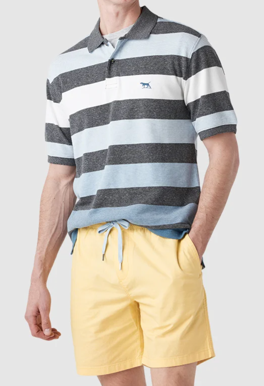 A person in a Rodd & Gunn Cox Road Polo, short-sleeve with wide black, white, and light blue stripes, paired with yellow drawstring shorts. The polo features an embroidered animal logo on the chest and is crafted from Australian cotton. Background is light gray.