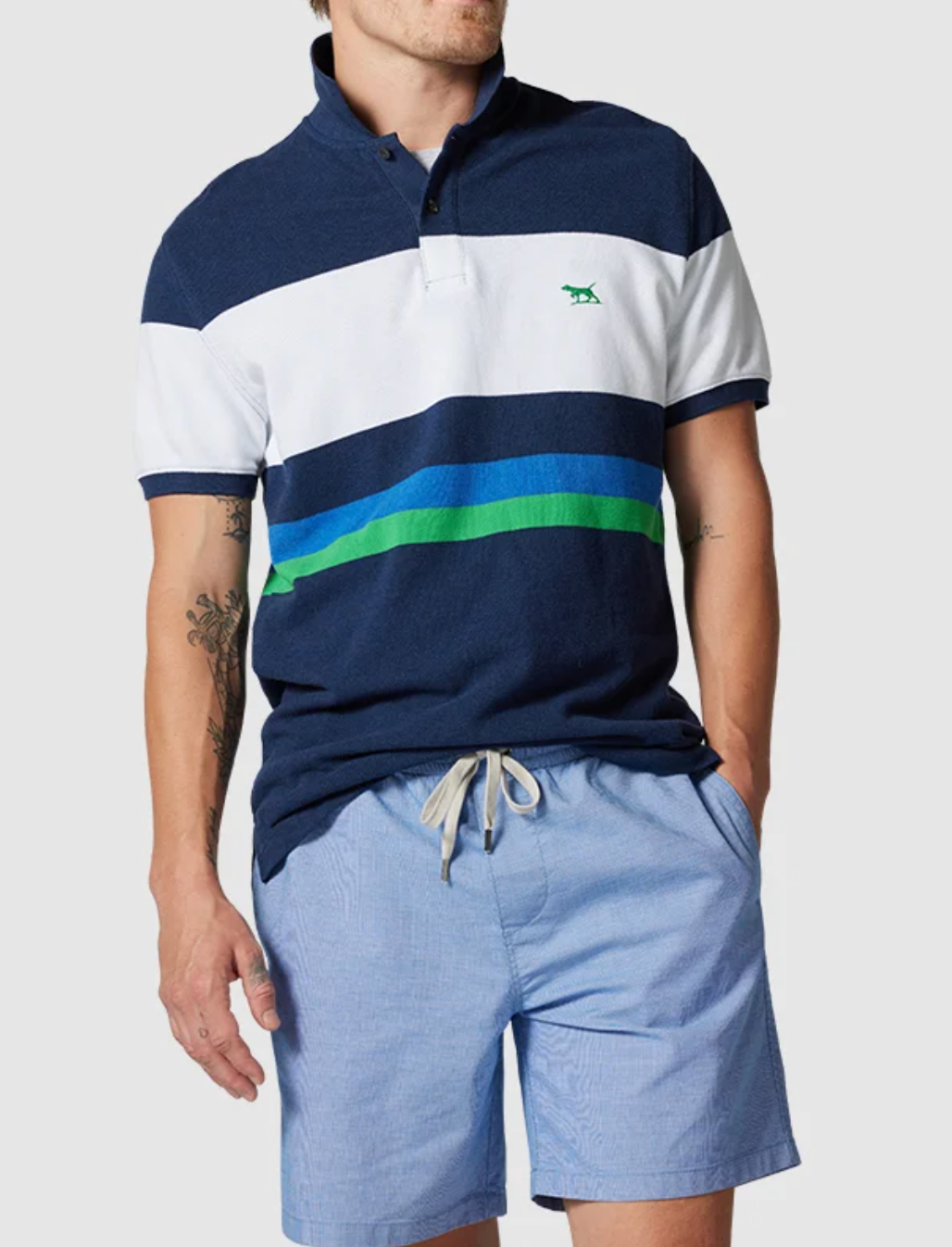 A man wears a Rodd & Gunn Willow Park Polo in navy, white, and blue stripes made from Australian Cotton with an embroidered chest logo. He pairs it with light blue drawstring shorts and has his left hand in the pocket against a plain light gray background.