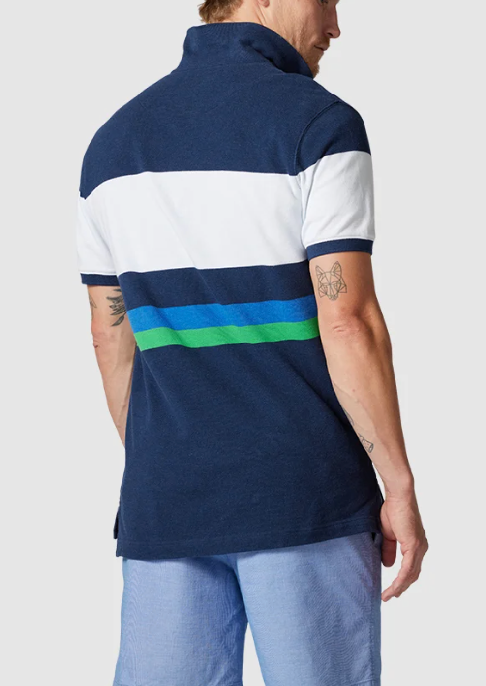 A person wearing the Rodd & Gunn Willow Park Polo, crafted from Australian Cotton with navy and green stripes, stands facing away. Their short hair highlights their arm tattoos, paired with light blue shorts for a relaxed look.