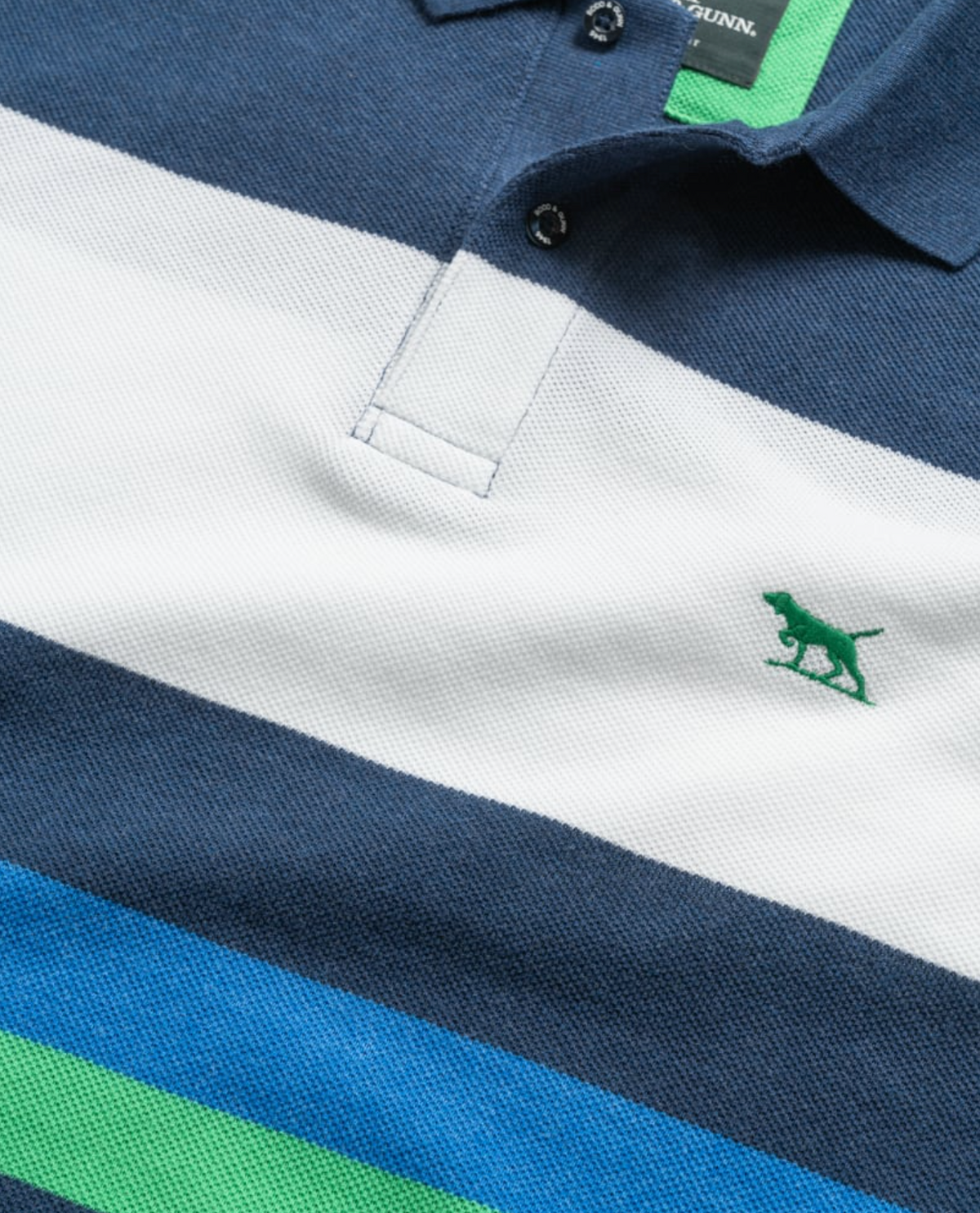 Close-up of the Rodd & Gunn Willow Park Polo, a sports fit shirt with navy, white, green, and light blue stripes. Made from soft Australian cotton, it features a green embroidered dog logo on the chest and a partially visible buttoned collar.