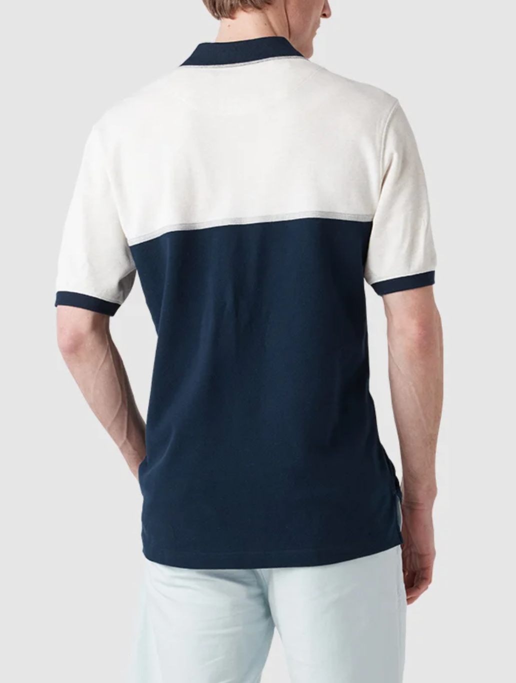 A man is wearing a Rodd & Gunn Castleford Polo made from Australian cotton, featuring a white upper half and navy blue lower half with dark blue trim on the collar and sleeves. He complements it with light blue pants and turns away, highlighting the stylish back of the ensemble.
