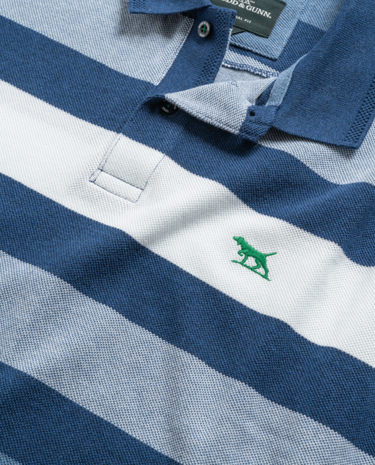 Close-up of a Rodd & Gunn Spencerville Polo, made from Australian cotton, showcasing blue and white stripes with a small green dog logo embroidered on the chest. The polo has a blue collar, with the top button undone, revealing the brand tag inside.