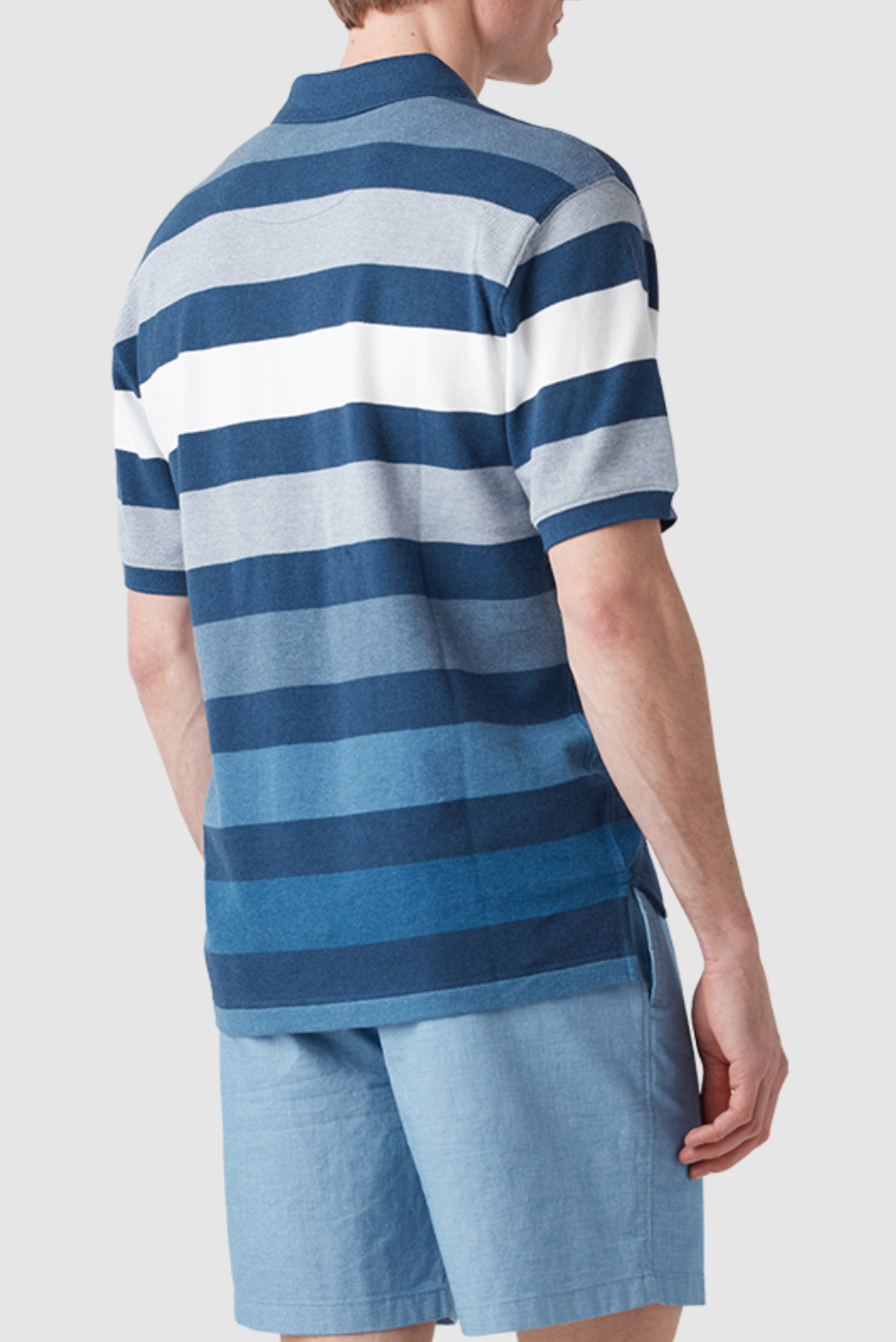 A person in a Rodd & Gunn Spencerville Polo, featuring blue and white horizontal stripes and made from Australian Cotton, pairs it with light blue shorts. They stand against a plain light gray backdrop, subtly displaying the embroidered logo while facing away from the camera.