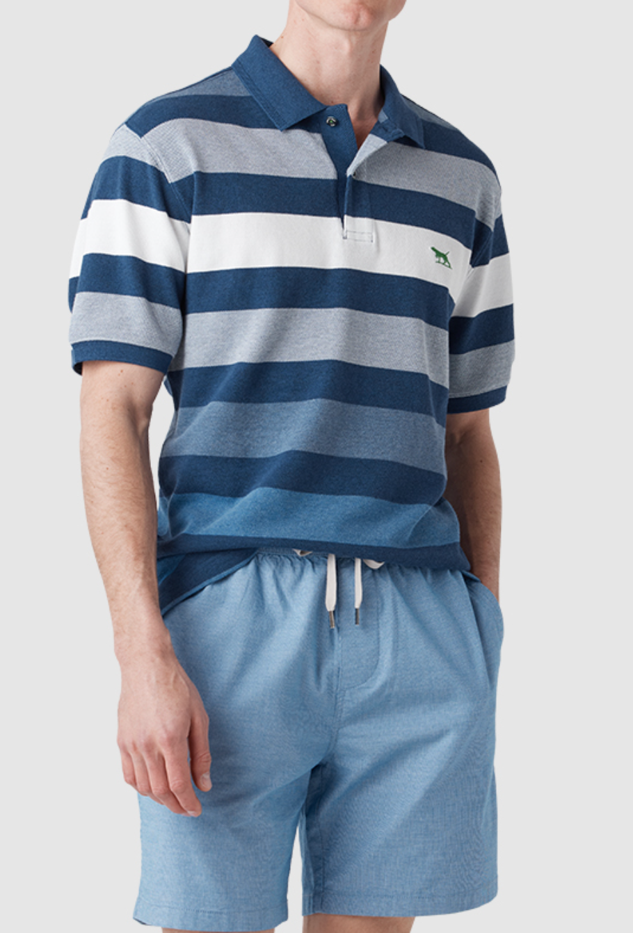 Someone is wearing a Rodd & Gunn Spencerville Polo with blue and white stripes and an embroidered chest logo, paired with light blue shorts. Their hands rest in the shorts' pockets against a plain light gray background.