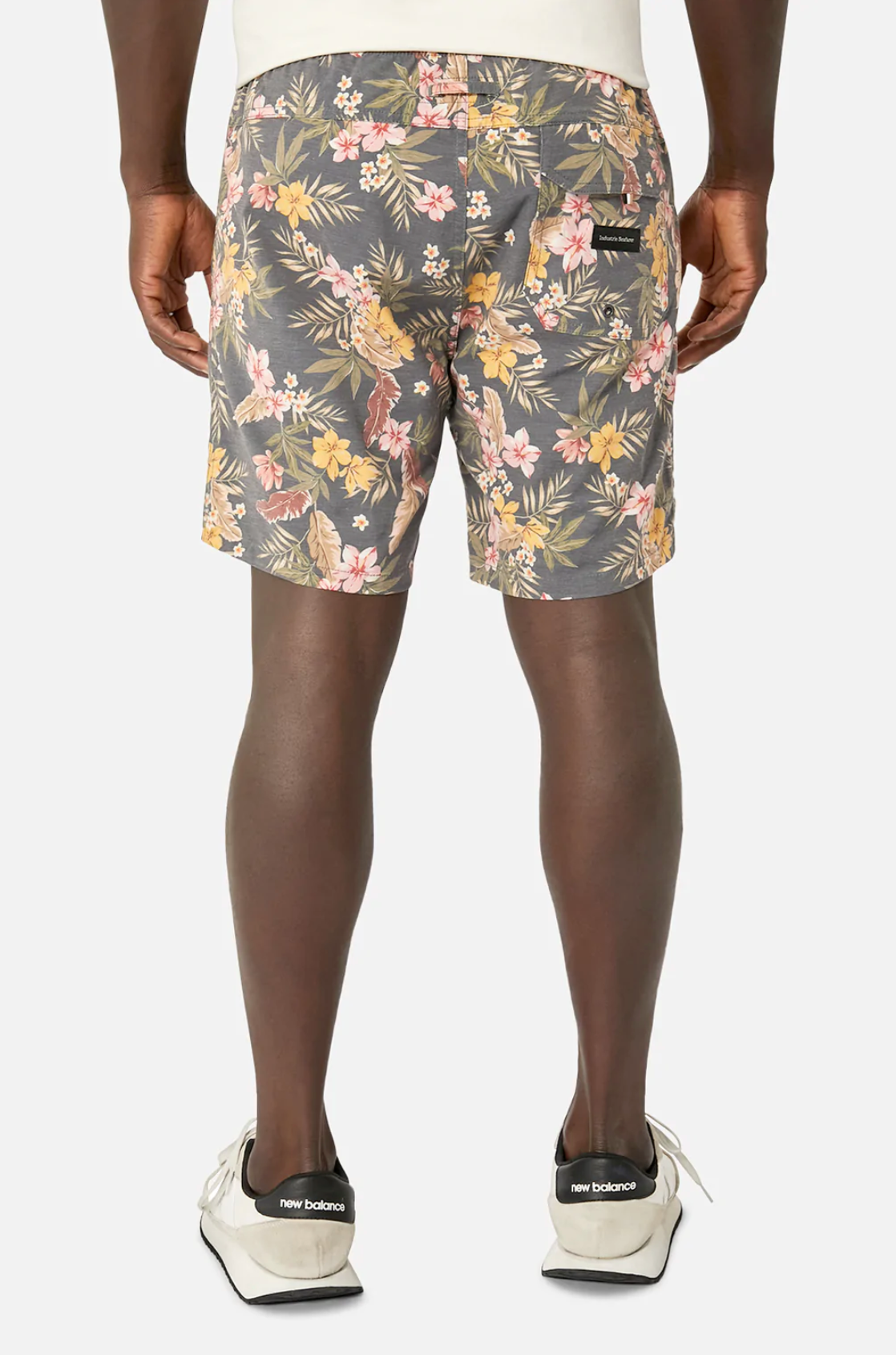A person is seen from the back wearing Industrie The Livadi Bahama Short with a floral pink and yellow design. They are also wearing a white shirt and gray New Balance sneakers with white soles. The shorts feature an elasticised waist for added comfort against a plain white background.
