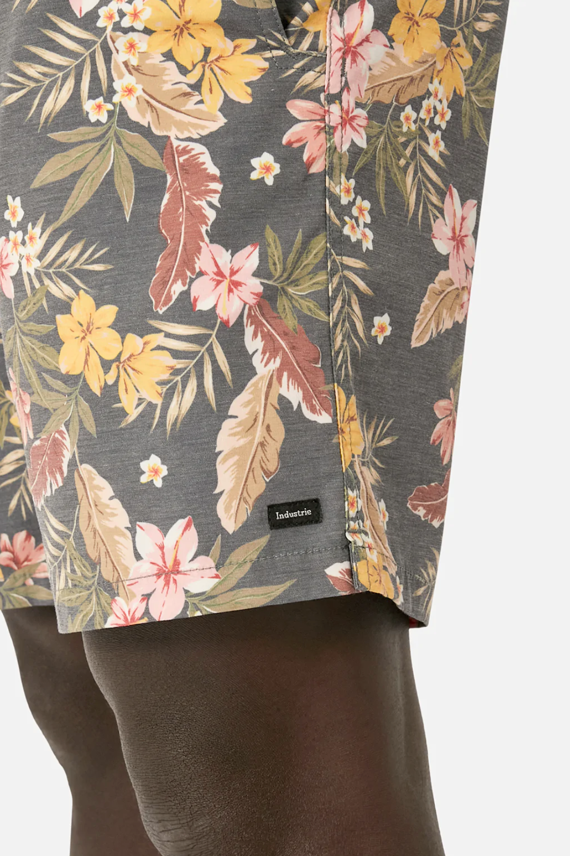 A close-up shows Industrie The Livadi Bahama Short, with floral patterns of yellow, pink, and brown on gray. A small black Industrie tag is visible near the hem. These board shorts feature an elasticized waist and are worn by a person.