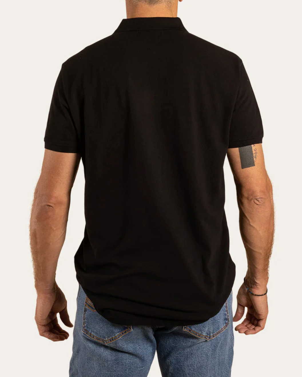 The rear view shows a person in a Ringers Western Essential Polo, black and short-sleeved, made of 100% cotton, paired with blue jeans. A tattoo decorates their left upper arm against a plain, light-colored background.