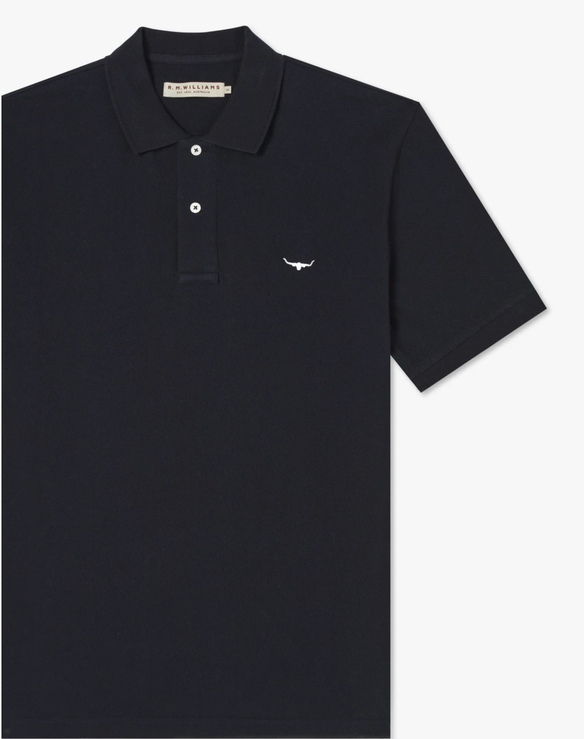 The RM Williams Rod Polo is a black cotton pique polo shirt with a folded collar, featuring a small white longhorn embroidery on the left chest. It has short sleeves, R.M.Williams branded buttons on the three-button placket, and a beige tag with branding information inside the collar.