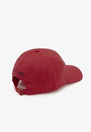 The RM Williams Mini Longhorn Cap, in a classic red twill design, is viewed from the back, highlighting its adjustable strap with a metal buckle. "EST. 1842" is embroidered in black near the adjustment strap against a plain white background.