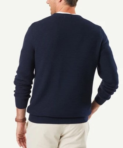 A person stands with their back to the camera, wearing the Gazman Textured Cotton Crew in navy blue and beige pants. Their right hand is placed on their waist. The comfortable jumper, made from combed cotton, features a ribbed texture, while the person's left hand rests down by their side.