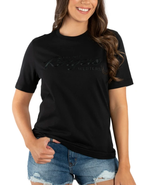 A woman in a Ringers Western Womens Columbia Emboss Logo T-Shirt and denim shorts radiates casual elegance. Her long, wavy hair and slight smile add confidence to her laid-back vibe, with a hand casually in her pocket.