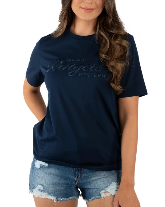 A woman wears the Ringers Western Womens Columbia Emboss Logo T-Shirt made of 100% cotton in navy, featuring subtle "Established 2012 Ringers Western" embossed lettering. She pairs it perfectly with denim shorts and has long, wavy hair.