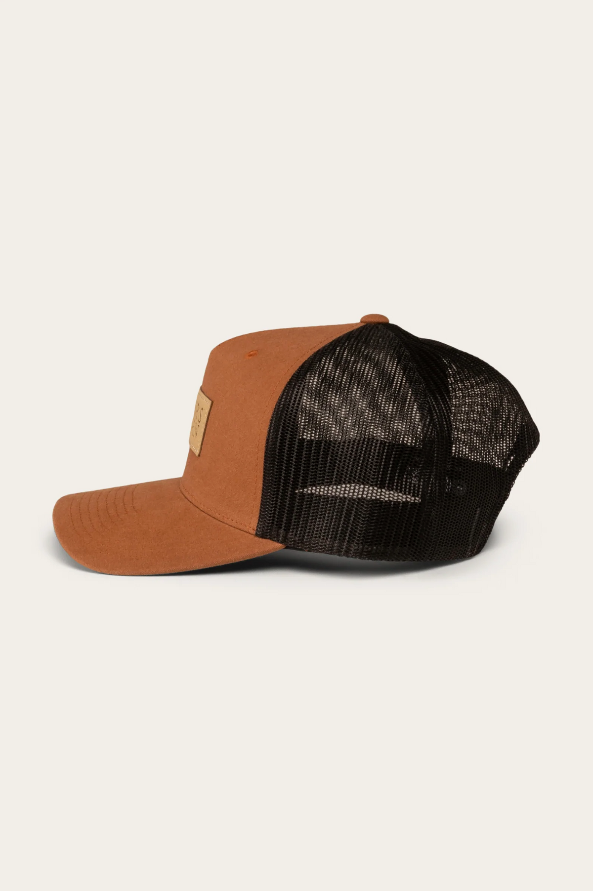 The Ringers Western Dustin Trucker Cap features a side view design with a brown cotton twill front panel and brim, black mesh back, and is adorned with a small rectangular patch on the front. The backdrop is light gray.