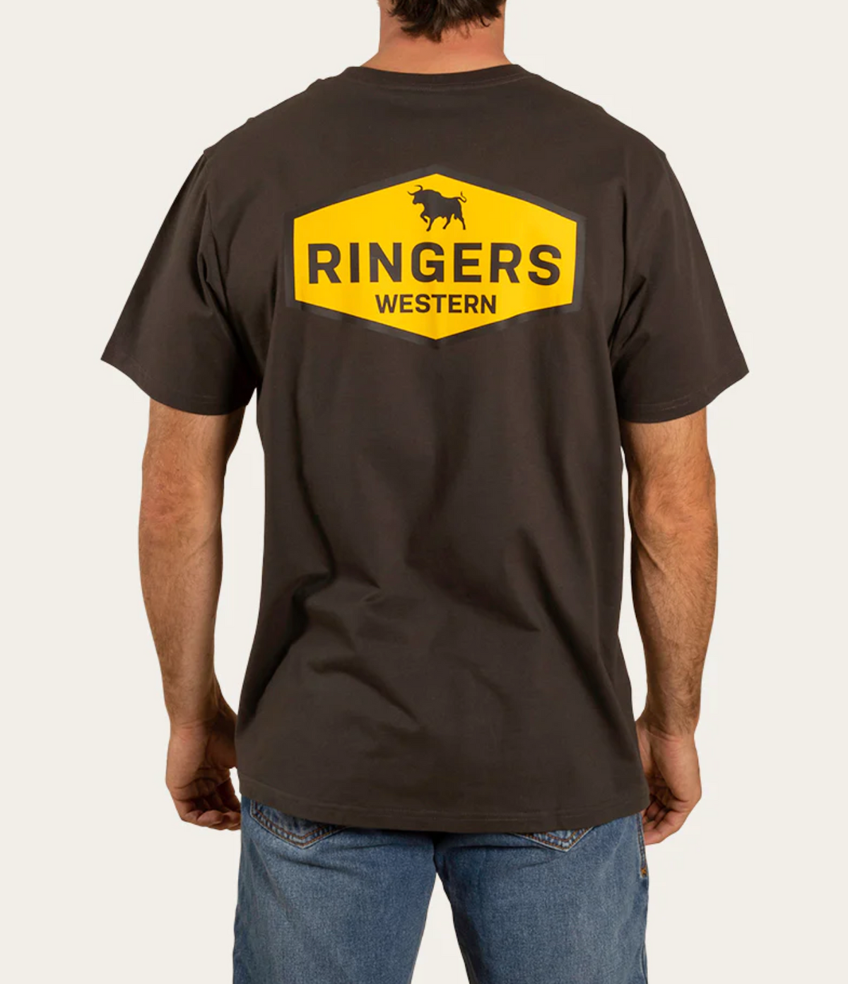 A person wearing a Ringers Western Servo Loose Fit T-Shirt in dark color with a yellow hexagonal logo on the back, featuring a bull silhouette and "RINGERS WESTERN" text. They pair it with blue jeans.