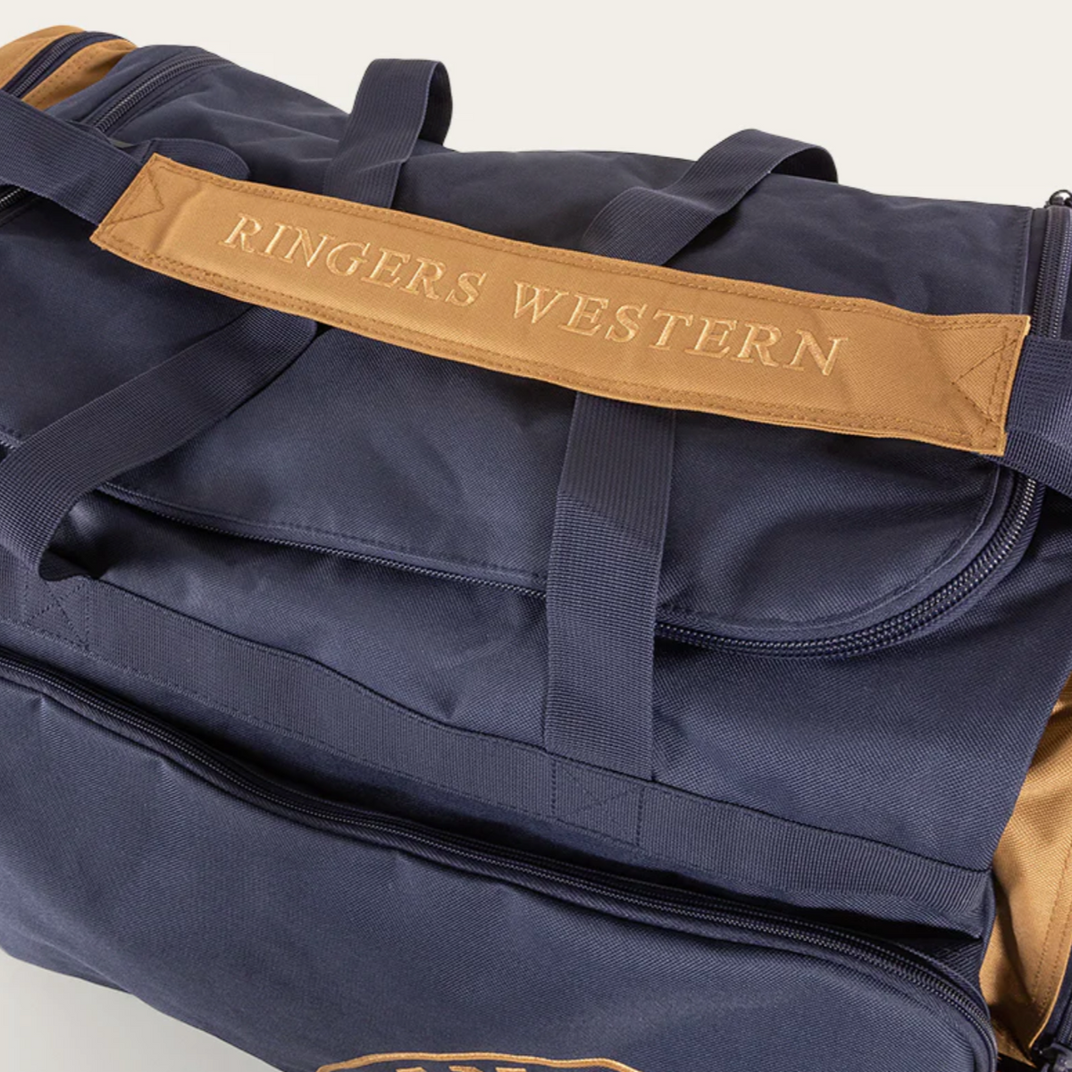 A close-up of a dark blue Ringers Western Rider Sports Bag featuring brown accents and handles. The bag proudly displays the brand name "Ringers Western" embroidered in gold on a brown strap at the top. Visible zippers and compartments highlight its practical design.