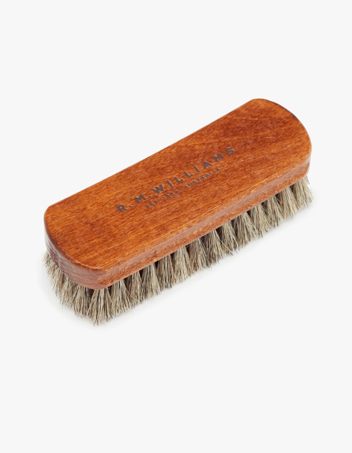 The RM Williams Medium Brush is a rectangular wooden leather boot brush featuring light brown horse hair bristles. The top of the brush is engraved with "R.M. Williams" and "Made in England." The densely packed bristles are ideal for polishing shoes.