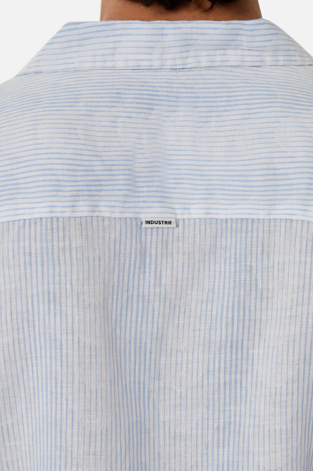 The back view of The Tropicana Shirt by Industrie in light blue and white stripes features a label on the collar. Made from 100% linen, its thin and lightweight fabric offers a casual and comfortable style with a relaxed fit.