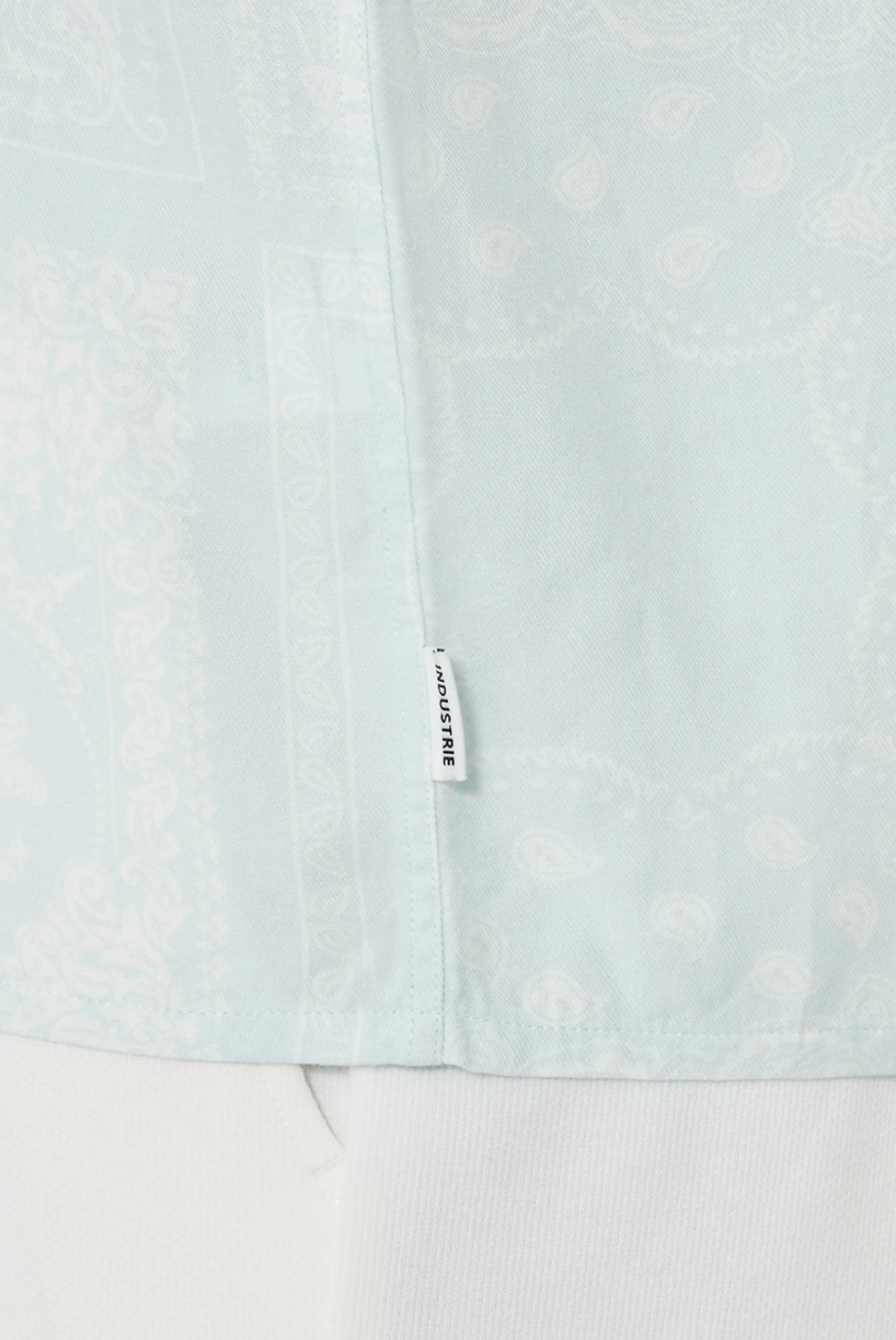 Close-up of a light blue fabric with a subtle paisley and geometric print. A small white tag with black text reads "JACQUEMUS," but the product is actually the Industrie The Stryder Shirt, a relaxed fit button-down. The bottom of the image shows a hint of white fabric.