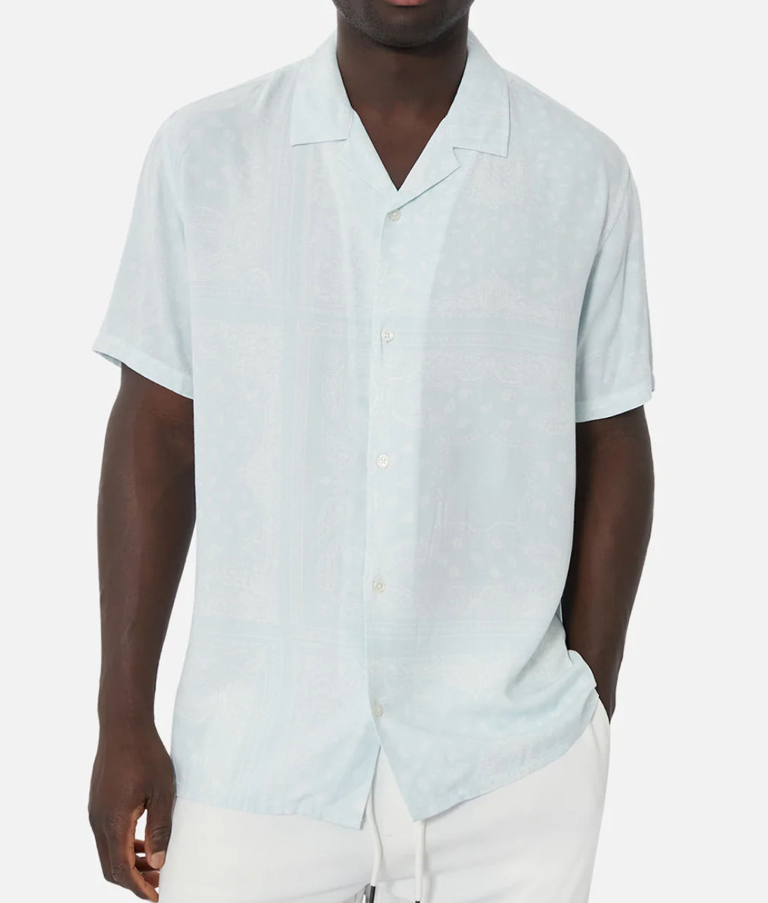 A person in an Industrie The Stryder Shirt, light blue with subtle geometric patterns, stands against a white background. Their left hand is in their pocket and they pair it with relaxed-fit white pants.