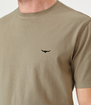 A close-up of a person wearing an olive green RM Williams Parson T-Shirt made from breathable cotton, featuring a small black bull logo with Longhorn embroidery on the left chest. The person's head is partially out of frame.