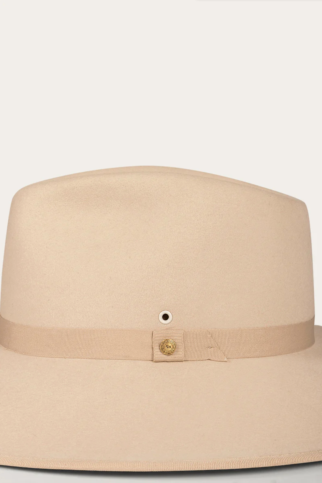 The Ringers Western Woodford Hat, a premium felt fedora, showcases a wide flat brim and a beige grosgrain ribbon with a small gold button detail, exuding timeless style against its pristine white background.