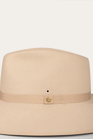 The Ringers Western Woodford Hat, a premium felt fedora, showcases a wide flat brim and a beige grosgrain ribbon with a small gold button detail, exuding timeless style against its pristine white background.