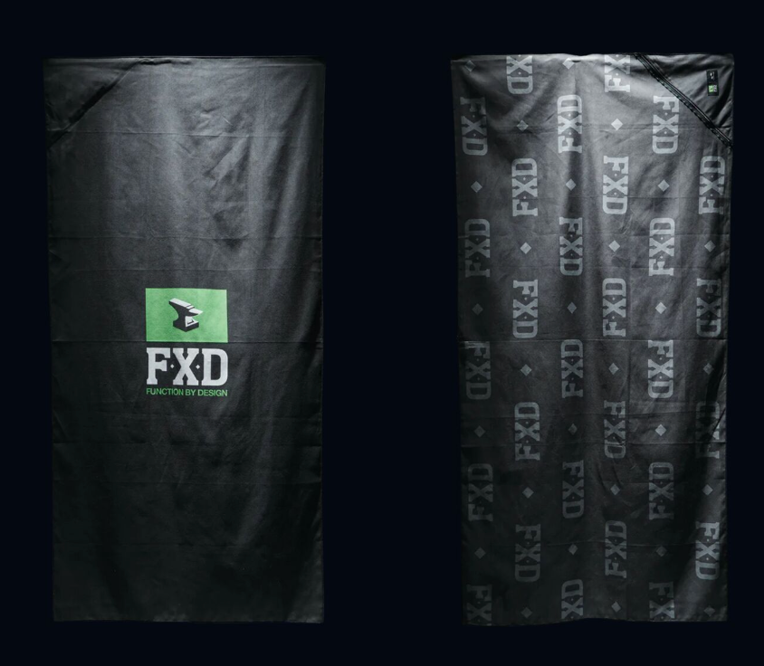 Two black FXD WAT-1 X Slowtide Work Towels are shown against a dark background. The left towel has a green "FXD" logo with "FUNCTION BY DESIGN," made from recycled polyester, while the right towel displays a gray fast-drying repeated "FXD" pattern.