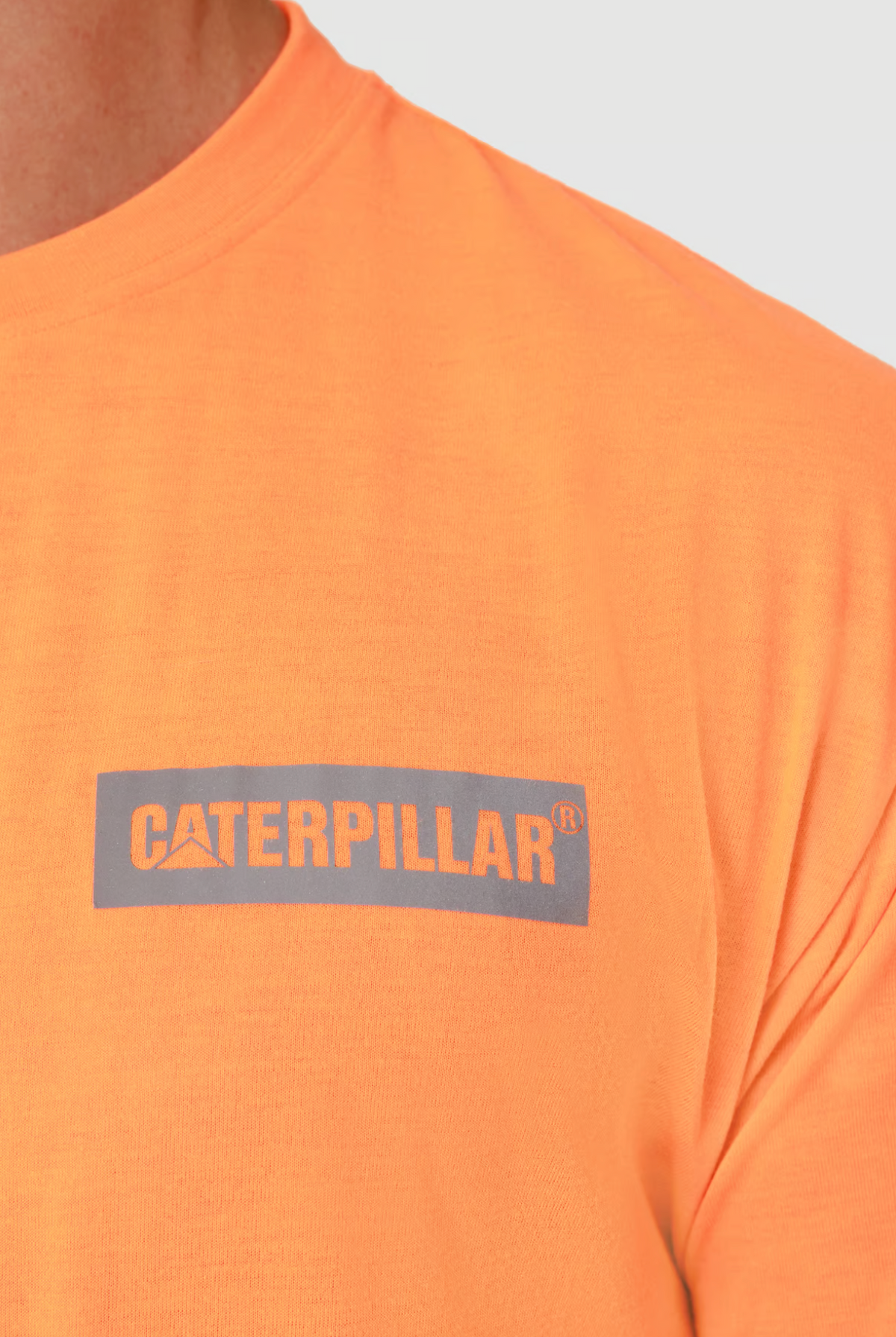 A close-up shows a person in the CAT Triton Block Tee, an orange 100% cotton shirt with the "CATERPILLAR" logo in gray on the chest. Its comfortable fit and neutral design make it ideal for any occasion.