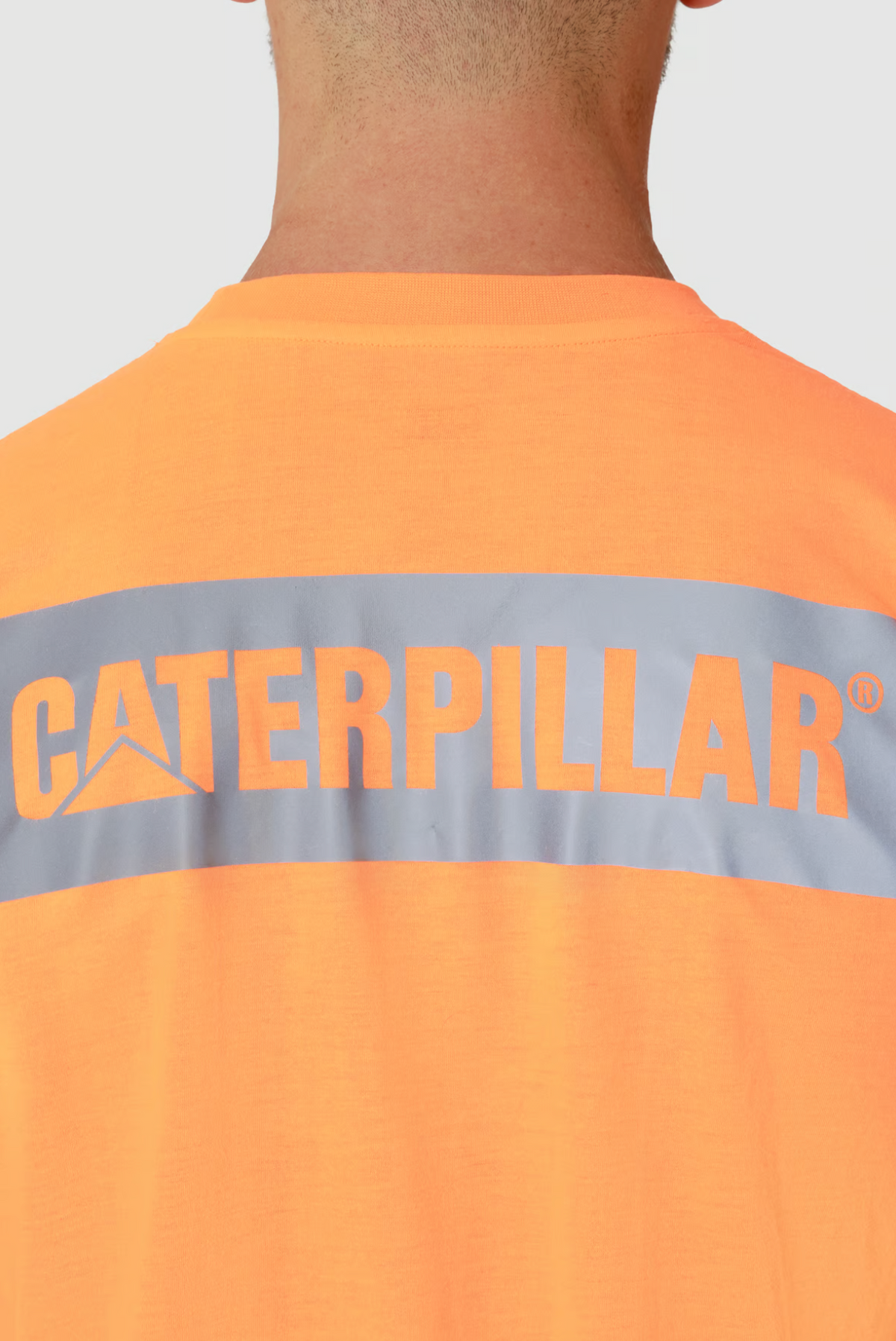 The image displays the back of a person wearing the CAT Triton Block Tee, featuring an orange tee with a gray stripe and "Caterpillar" in bold orange text. Made from 100% cotton, it suits their short hair while they face away from the camera.