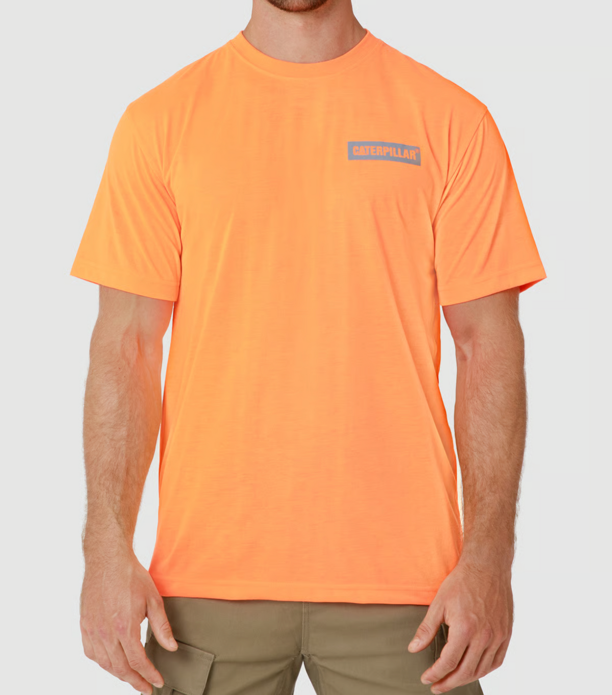 A person wearing the CAT Triton Block Tee, an orange 100% cotton T-shirt with the Caterpillar logo on the chest, paired with beige cargo pants. The light gray background enhances the tee's vibrant hue.