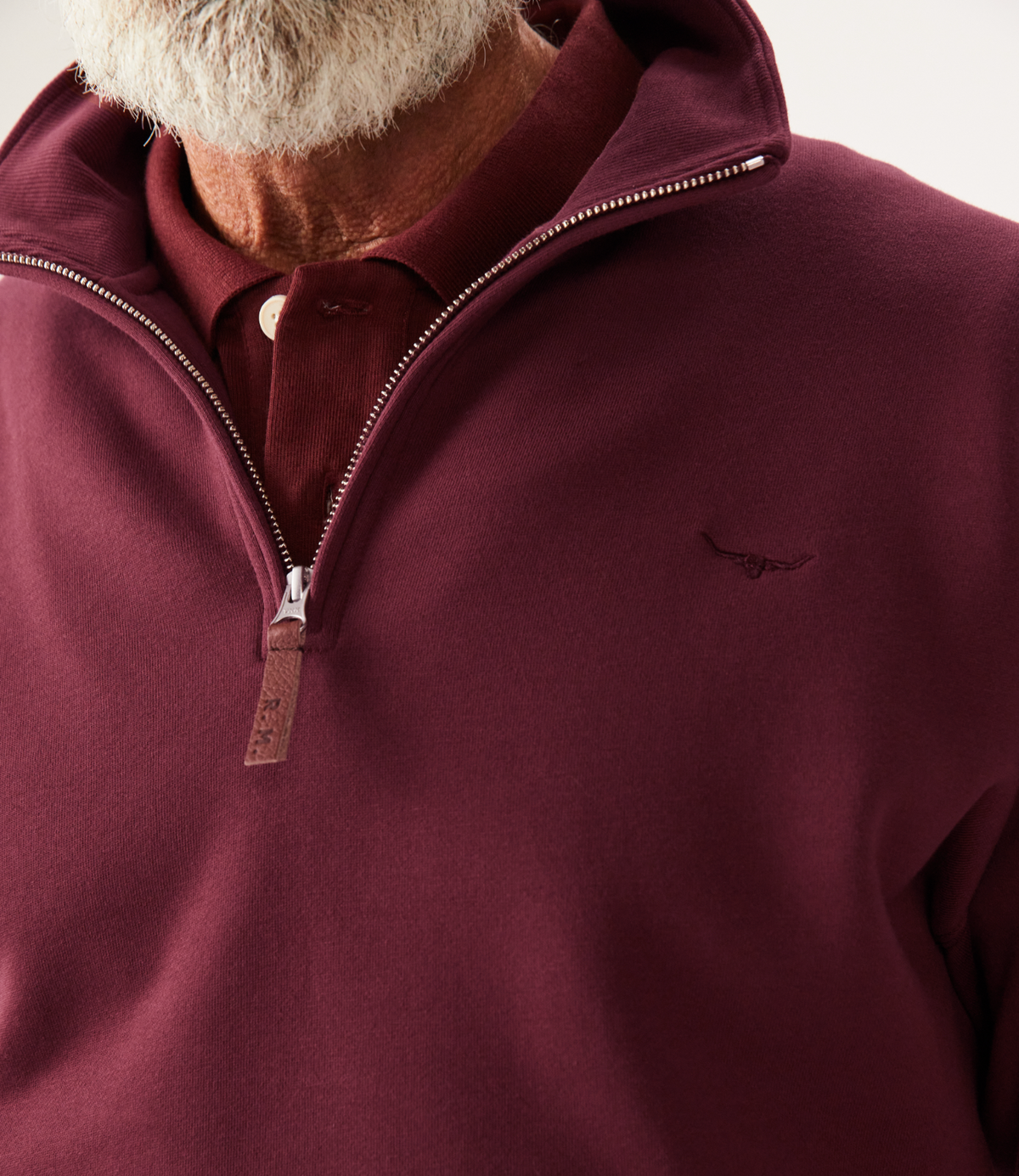 A man with a white beard wears an RM Williams Mulyungarie Fleece quarter-zip sweatshirt in maroon over a matching polo. The brushed cotton fleece has a small embroidered chest logo. Only his lower face and upper torso are visible.