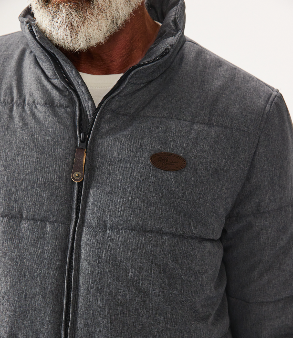 Close-up of a man with a gray beard wearing the RM Williams Patterson Creek Jacket, a quilted puffer with zip-up collar and brown logo patch on the chest, over a white shirt. Water and winter resistant for added comfort.