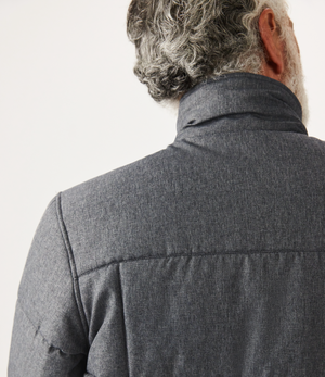 A person with curly gray hair and a beard is seen from behind in an RM Williams Patterson Creek Jacket, dark gray with a high collar. The quilted, textured fabric adds depth, set against a plain off-white background that highlights the jacket's understated elegance.
